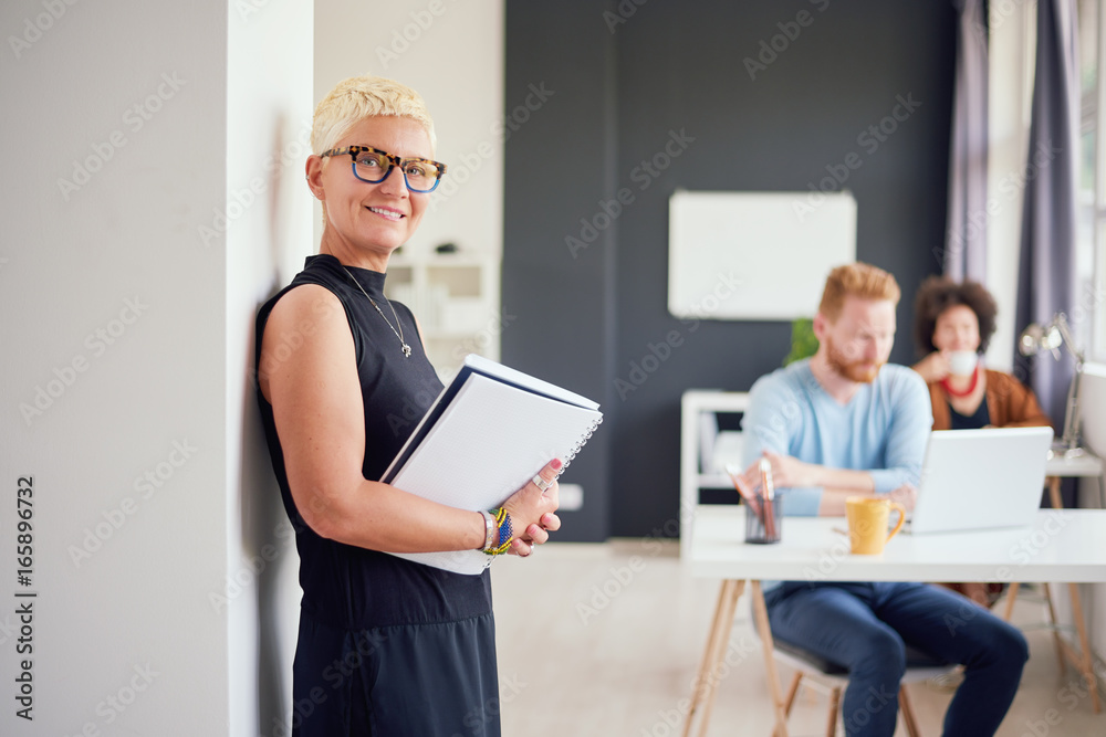 Senior standing in modern office against the wall and holding notebook
