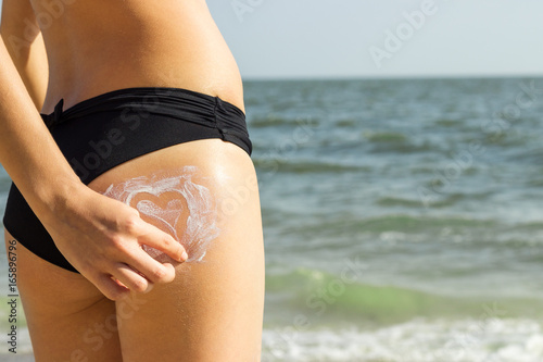 Attractive woman with healthy skin applying sunscreen in the form of heart on her buttocks