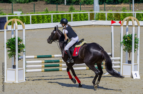 Young girl jumping with black horse. Blond pretty little girl going jump a hurdle in a competition. Girl with crow horse during equestrian showjumping. Bright sunny day.