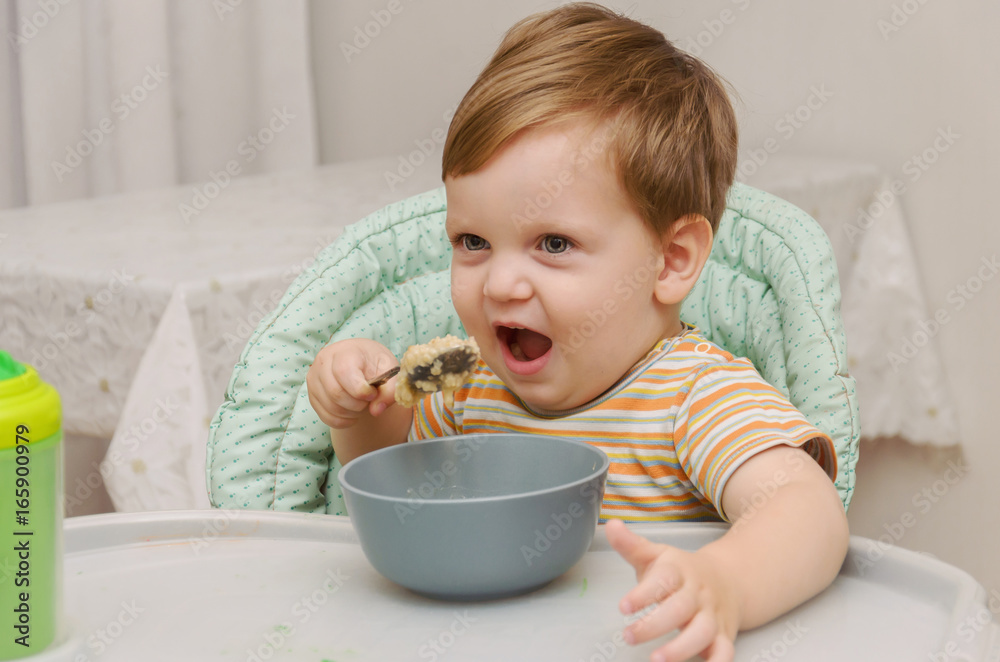 little blond boy in a striped T-shirt eats porridge with a spoon from a gray plate little blond boy in a striped T-shirt eats porridge with a spoon from a gray plate