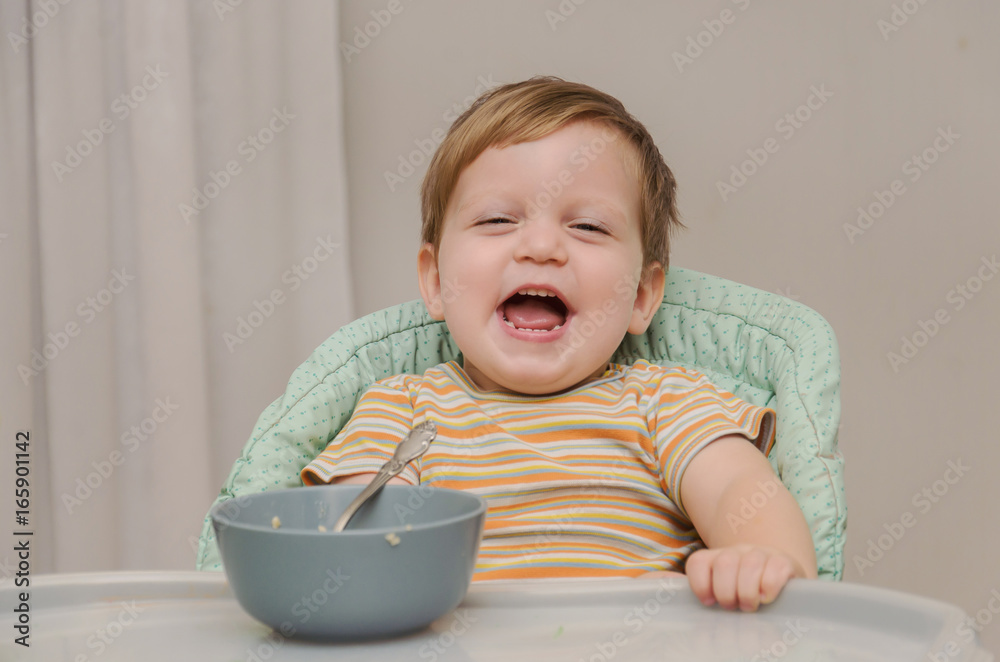 little blond boy in a striped T-shirt eats porridge with a spoon from a gray plate little blond boy in a striped T-shirt eats porridge with a spoon from a gray plate