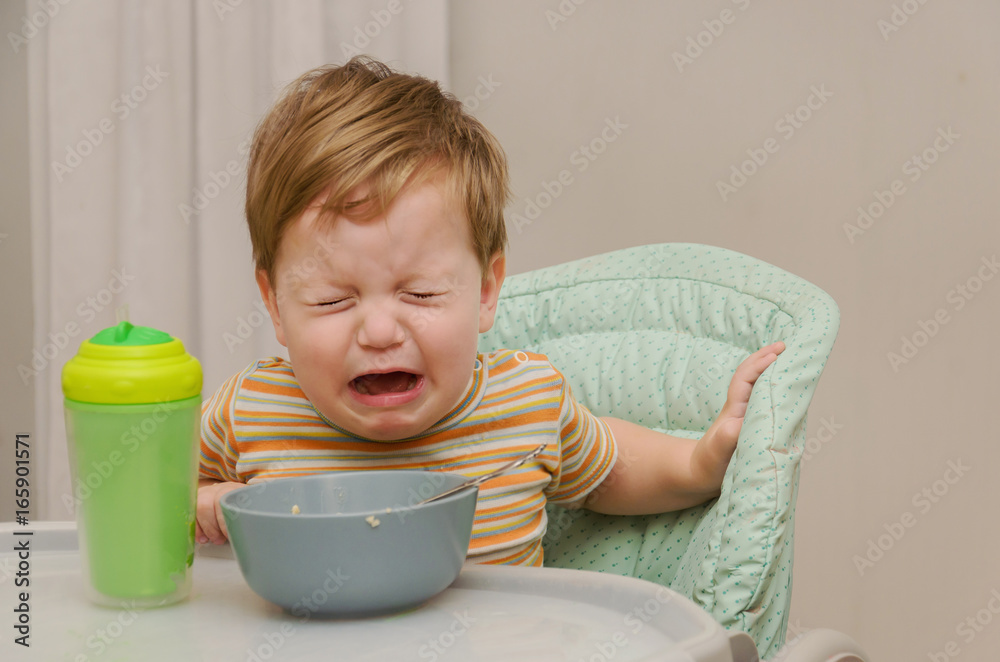 little blond boy in a striped T-shirt does not want to eat a porridge with a spoon from a gray plate. little blond boy in a striped T-shirt does not want to eat a porridge with a spoon from a gray plate.