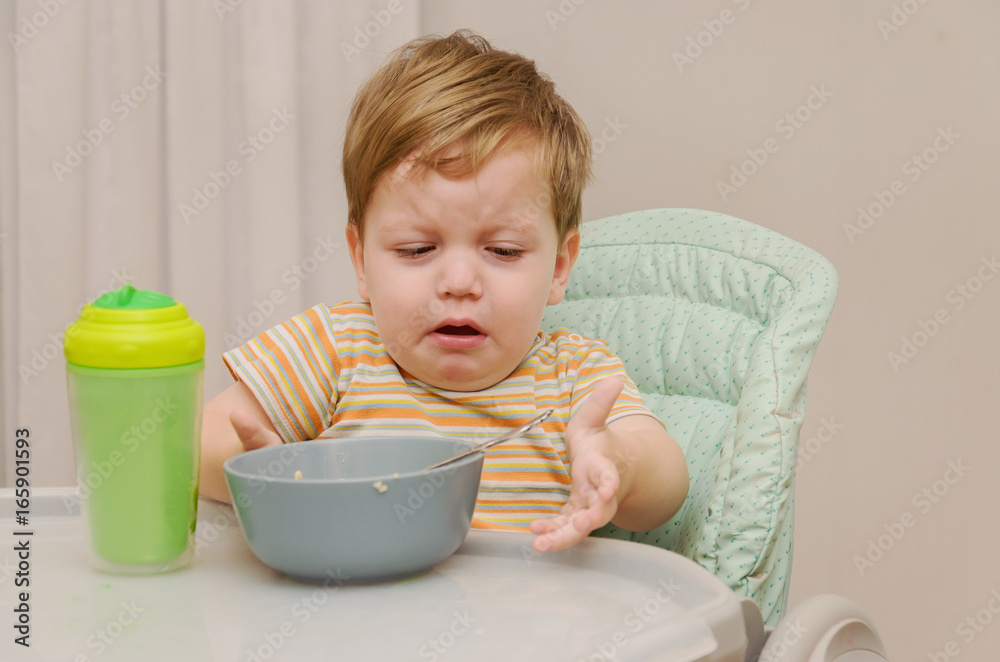 little blond boy in a striped T-shirt does not want to eat a porridge with a spoon from a gray plate. little blond boy in a striped T-shirt does not want to eat a porridge with a spoon from a gray plate.