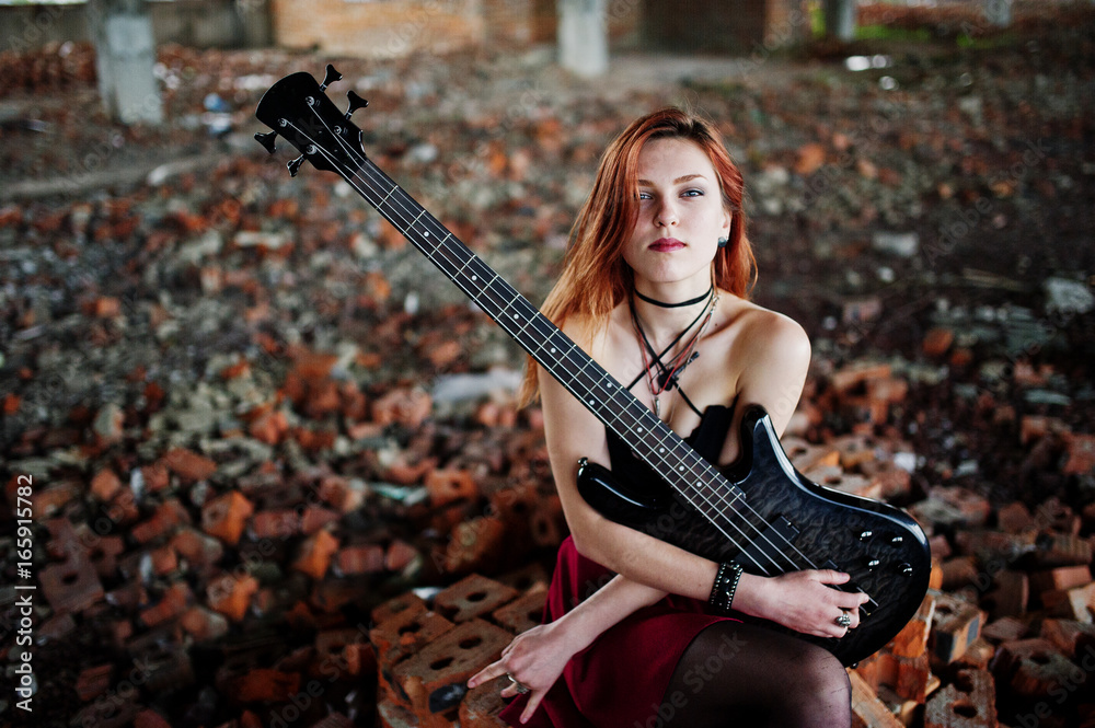 Red haired punk girl wear on black and red skirt, with bass guitar at abadoned place. Portrait of gothic woman musician.