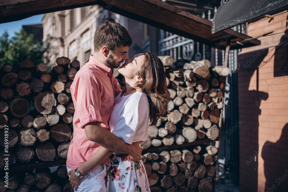 Stylish man and happy woman embrace in light on background of wooden firewood wall. Space for text. Happy couple are hagging, romantic moment.