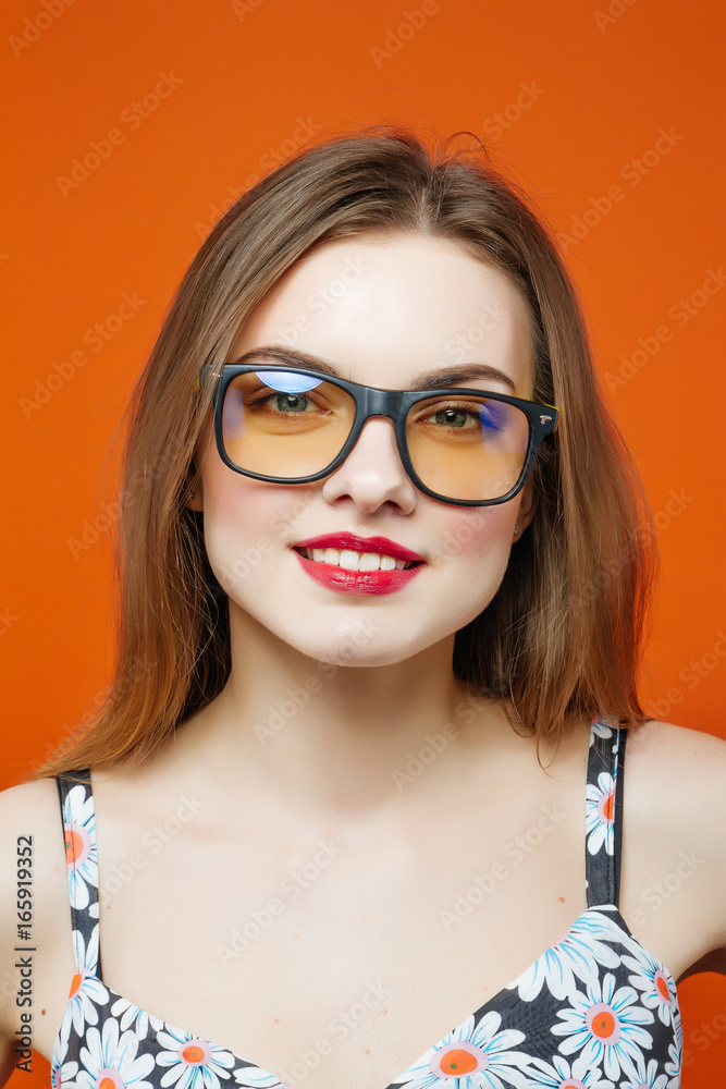 Portrait of Beautiful Female Model in Eyeglasses on Orange Background. Attractive Brunette with Sensual Lips and Dress with Flowers is Posing in Studio.
