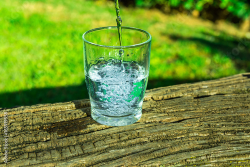 Process of pouring pure clear water into a glass from top, wooden log, green grass in the background, outdoors, health, hydration, cleansing, rejuvenation