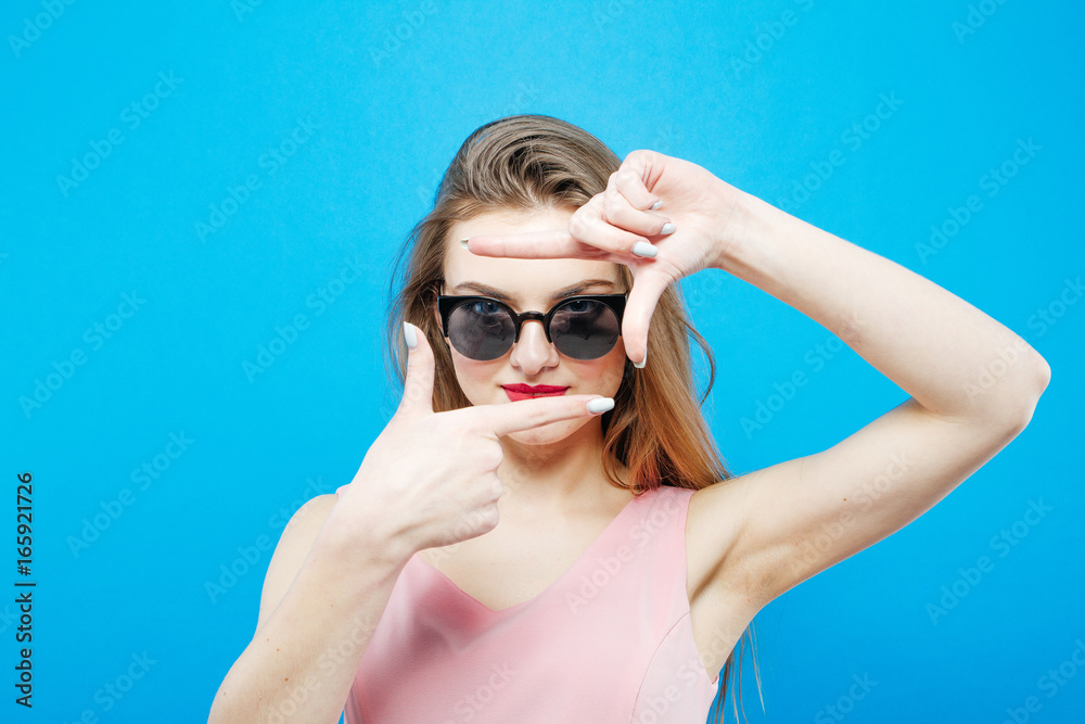 Amazing Brunette Model in Fashionable Sunglasses and Pink Dress is Having Fun in Studio on Blue Background.