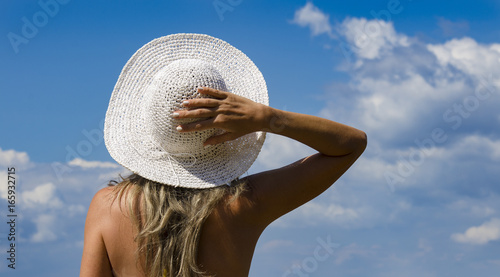 Young girl turned back, holding her hat with her hand behind her head, looking at a cloudy blue sky