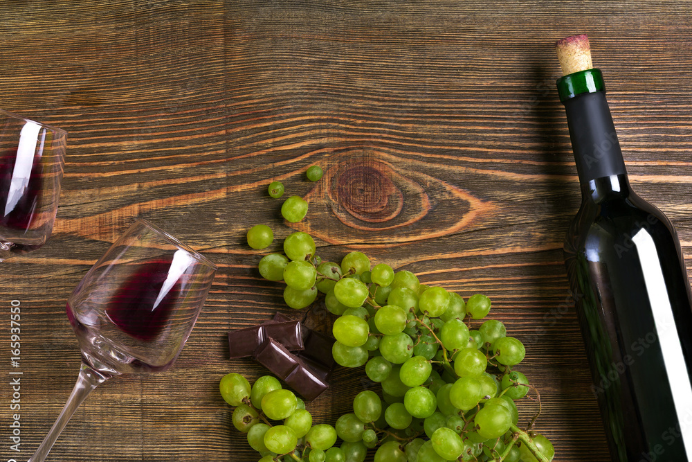 Obraz premium Red wine bottle, grape, chocolate and glasses over wooden table. Top view with copy space