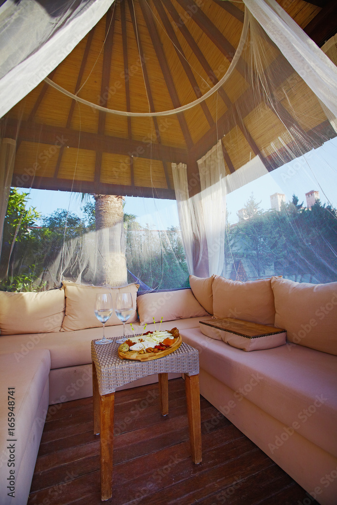 Fototapeta premium Gazebo on grass with soft pillows and sofa and served cheese and vine in resort place