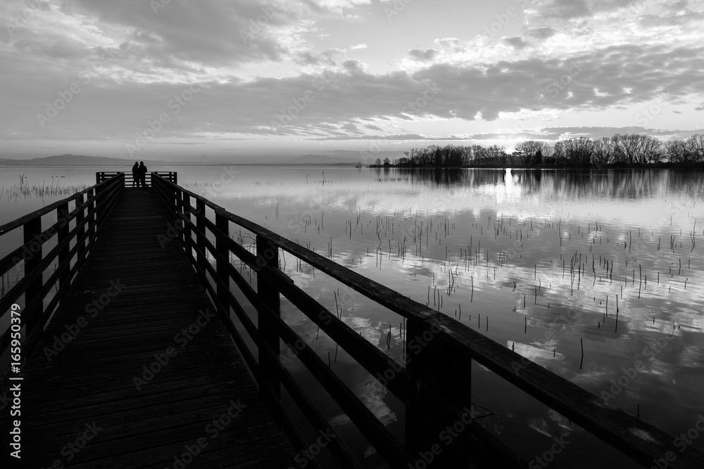 A first person view of a pier on a lake with two people on it and beautiful clouds and trees reflections on water