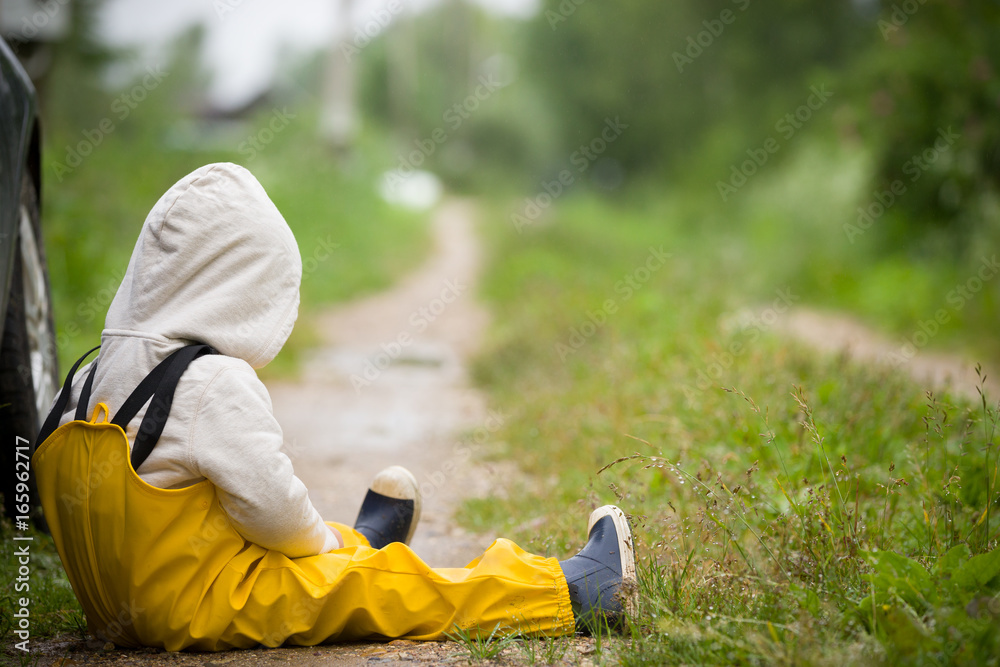 Portrait of small toddler boy in yellow rubber rain pants sitting on countryside road. Upset child. After rain. Outdoors Portrait of small toddler boy in yellow rubber rain pants sitting on countryside road. Upset child. After rain. Outdoors