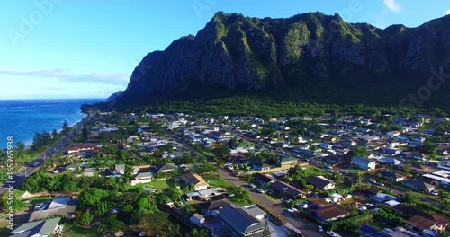 Highway Running Past Small Island Neighborhood in a Green Valley Surrounded by Imposing Green Mountains with Steep Cliff Walls - Aerial Footage on Oahu, Hawaii photo