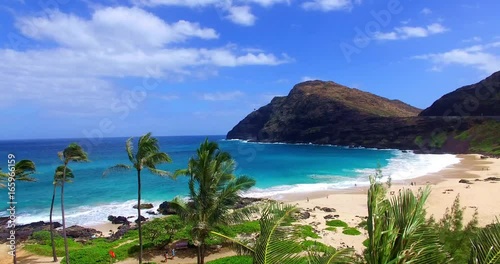 Blue Water Ocean in Sandy Cove with Beachgoers and Palm Trees Moving in the Wind Surrounded by Hills photo