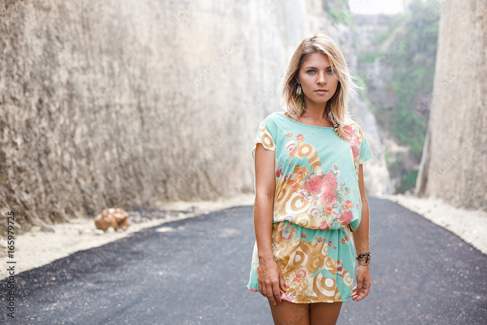Horizontal portrait of an attractive blonde girl in dress with flowers and abstract print and feather earrings is standing on the road between mountains
