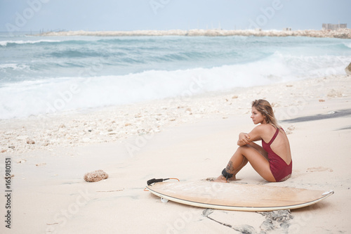 Attractive surf girl in dark-red swimming suit is sitting on the beach next to a surfboard in front of the ocean and looking away