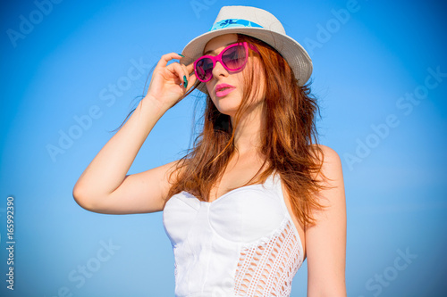 Girl with red hair on vacation in a white top, fedora hat and pink sunglasses against the blue sky. Life in pink and blue tones