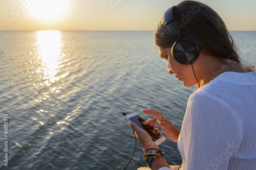 Young beautiful slim woman with long hair in jeans shorts and a white shirt sits on a wooden bridge with headphones and listens to music looking at the sunset by the sea 