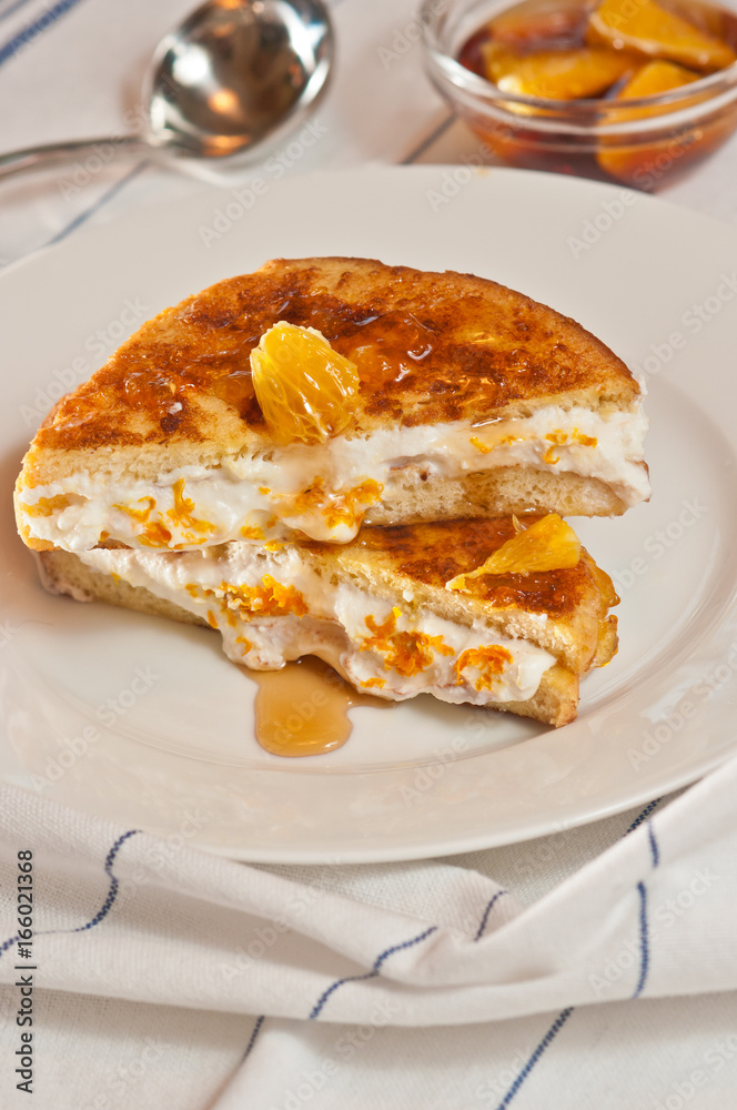 Baked orange cream french toast sandwich on a round, white plate with an artisan serving spoon and a clear glass bowl of orange sections on a whitetable cloth with blue stripes