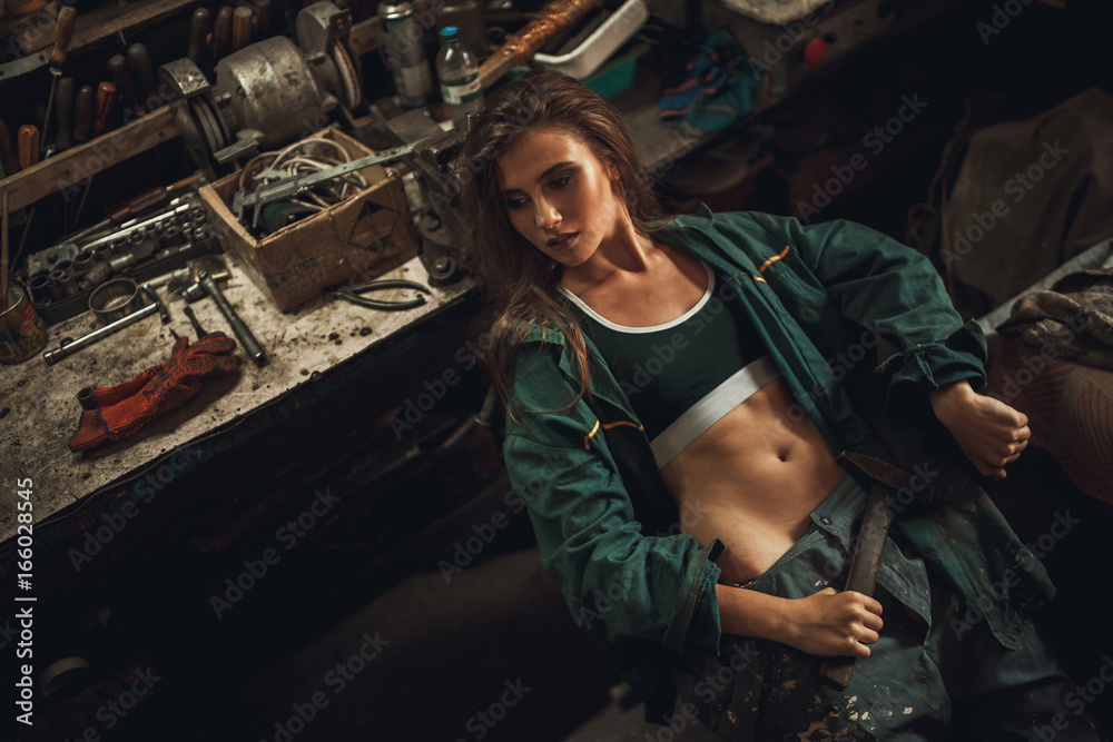 Girl worker in overalls sits in workshop with hammer in her hands.