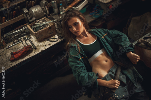 Girl worker in overalls sits in workshop with hammer in her hands. © Stanislav