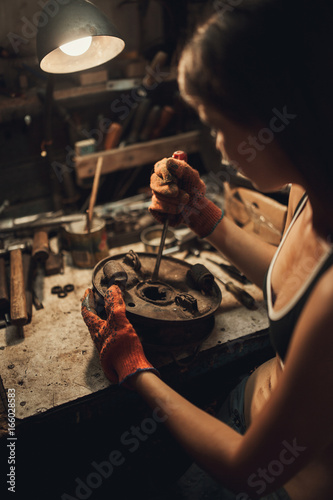 Girl worker in t-shirt sits in workshop among tools and unscrews spare part with screwdriver. Closeup.