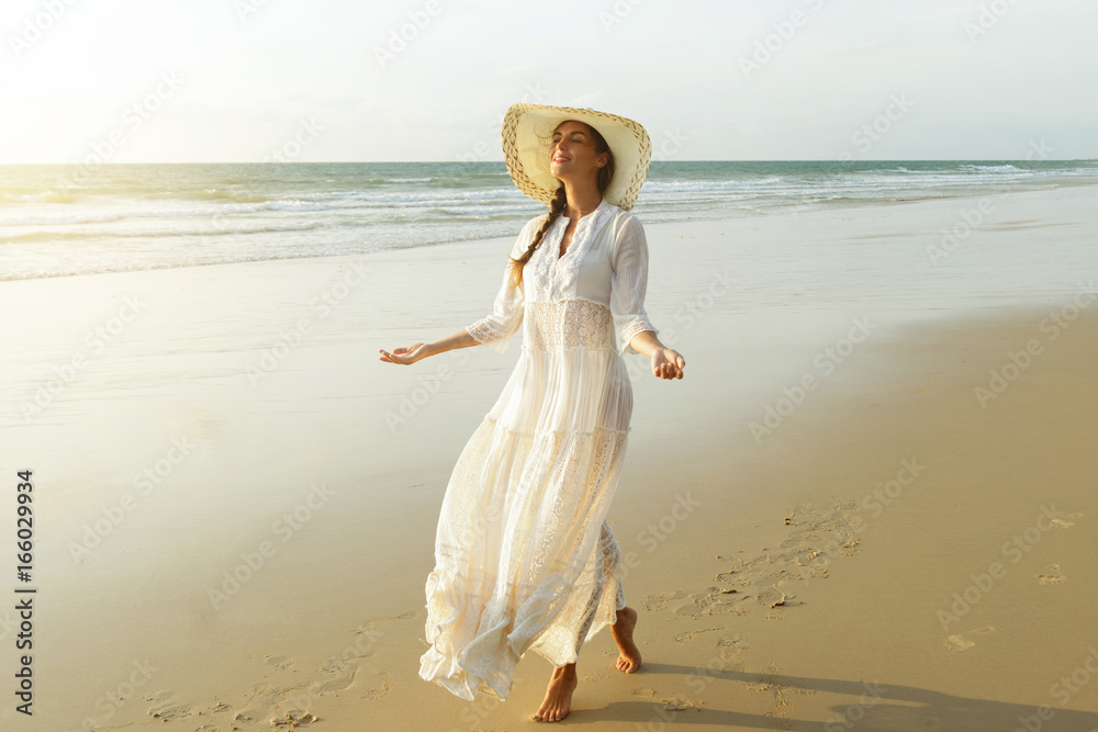 Woman wearing beautiful white dress is walking on the beach during sunset Woman wearing beautiful white dress is walking on the beach during sunset