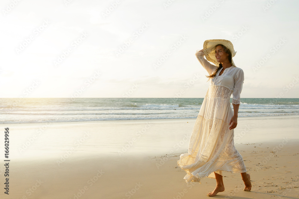 Woman wearing beautiful white dress is walking on the beach during sunset Woman wearing beautiful white dress is walking on the beach during sunset