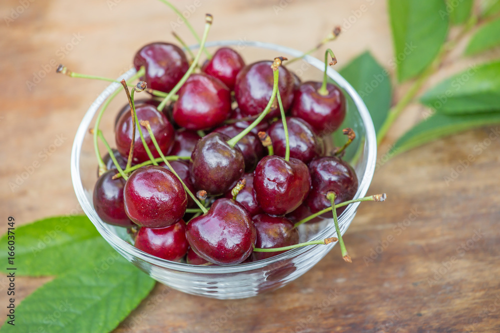 Ripe red cherries with green leaves in a bowl on wooden table closeup Ripe red cherries with green leaves in a bowl on wooden table closeup