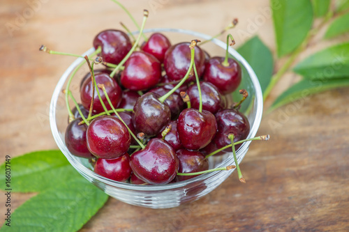 Ripe red cherries with green leaves in a bowl on wooden table closeup