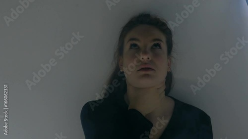 A young woman sitting behind a white wall chokes herself with one hand. Filmed from close up. photo