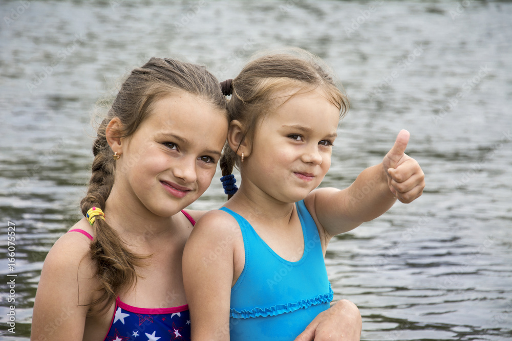 In the summer on the river, two small sisters in bathing suits are sitting in an embrace. In the summer on the river, two small sisters in bathing suits are sitting in an embrace.