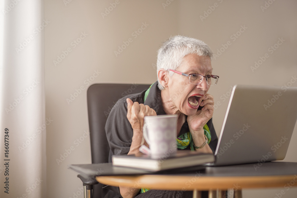 Senior old woman screaming at her laptop, enraged with news Senior old woman screaming at her laptop, enraged with news