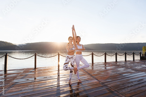 Two young women doing yoga at nature. Fitness, sport, yoga and healthy lifestyle concept - group of people making yoga pose on lake pier