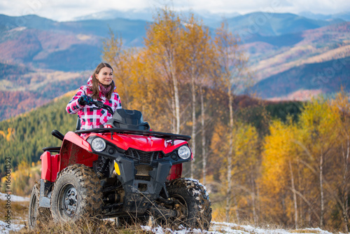 Brunette girl in winter clothes rides a quad bike on a snowy road with stunning autumn views of mountains, forests and trees with yellow leaves