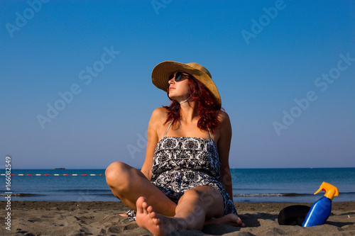 A young girl sitting on a beach in the sand, enjoying the sun and the sea, with a hat on her head and glasses