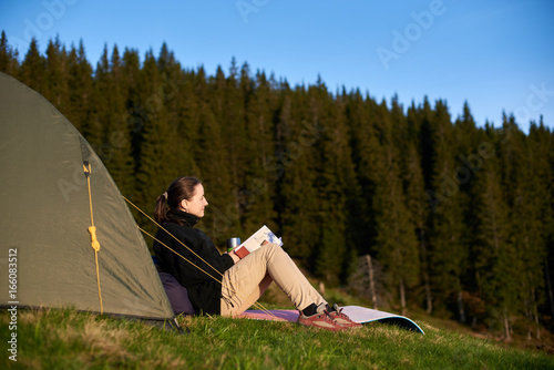 Young happy female hiker enjoying in the camping with book and cup of coffee near the tent in the evening