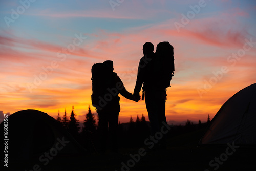 Silhouettes of a loving couple holding hands standing on top of the mountain while hiking together with their backpacks enjoying beautiful sunset with fiery colorful sky love relationships nature.