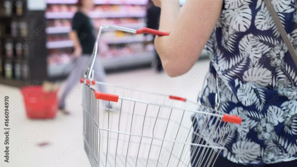 Closeup isolated shot of shop's iron handcart indoor. Woman's hand carry bag. Mistress slowly going between shelves and selecting the good in big modern marketplace