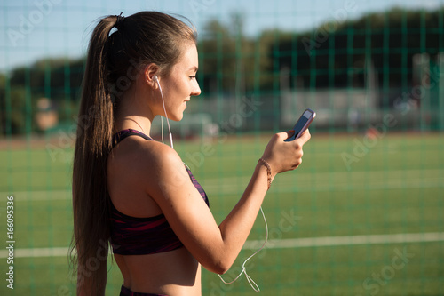 Young sportswoman in sportswear listen music with earphones and smart phone outdoors on stadium. Healthy lifestyle concept, modern technologies.
