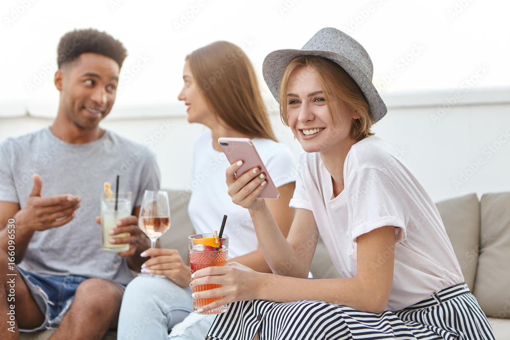 Friendly youngsters relaxing at restaurant, drinking cocktails or wine, communicating with each other. Cute woman in hat drinking mixed drinking typing message to her lover having joy while resting Friendly youngsters relaxing at restaurant, drinking cocktails or wine, communicating with each other. Cute woman in hat drinking mixed drinking typing message to her lover having joy while resting
