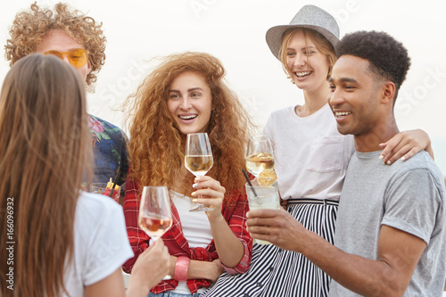 Diverse group of friends smiling as they cheers toast drinking wine and fresh cocktails embracing each other being glad of meeting. Cool party organised by youngsters. True friendship concept