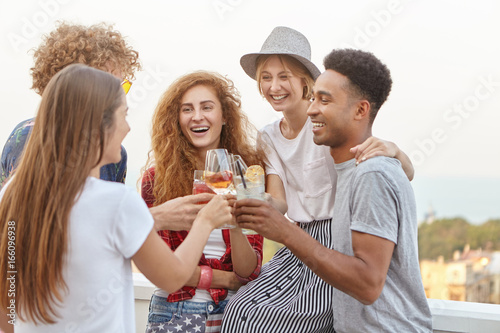 Party, celebration, rest and people concept. Interracial friends clinking drink glasses with happy expressions while standing at balcony celebrating somethng having party having fun together