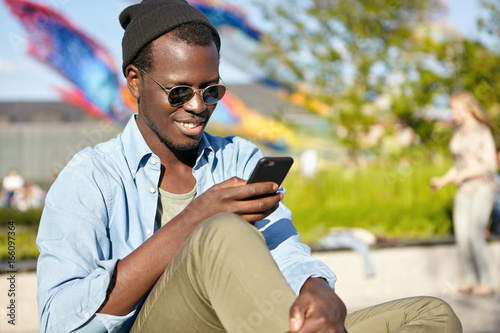 Valokuva Fashionable male with dark skin and bristle sitting outdoors checking his news feed or messaging via social networks using cell phone using free wi-fi