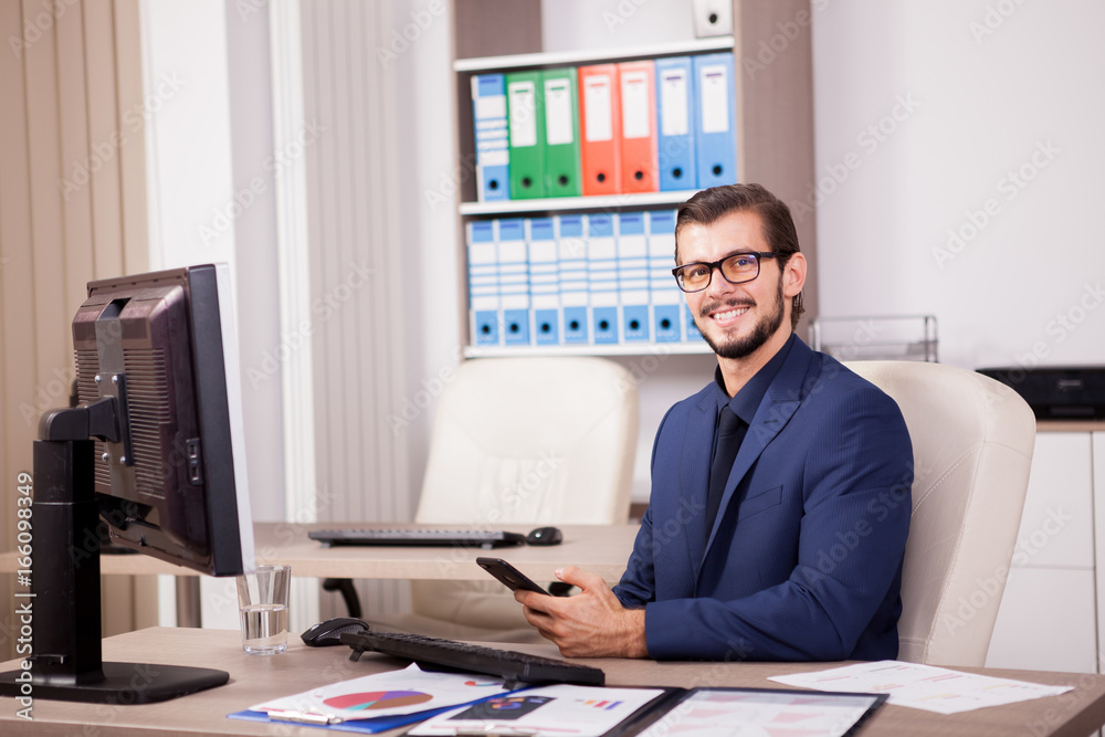 Smiling Manager with a smartphone in hands in his modern office. Image of successful businessman in blue suit. Communication and technology Smiling Manager with a smartphone in hands in his modern office. Image of successful businessman in blue suit. Communication and technology