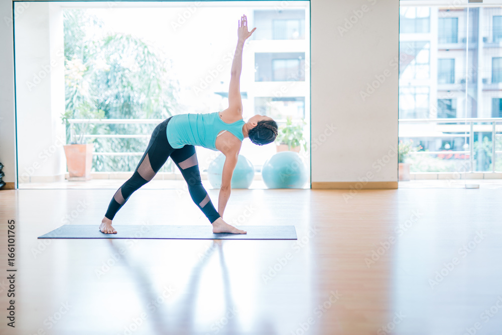 Portrait of beautiful young woman enjoying yoga indoors. Yogi girl working out in yoga studio twisting triangle Pose, Full length