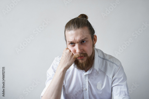 Sadness and boredom concept. Studio shot of unhappy young manager with hair knot and thick beard having upset and bored look, feeling tired and pissed off with boring working days at office