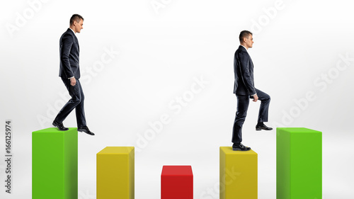 Two businessmen on white background stepping up and down green, yellow and green statistic columns.