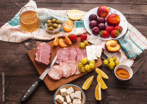 White wine and a set of meat delicacies, cheese and fruits on a dark, wooden background. Breakfast and cooking. View from above.